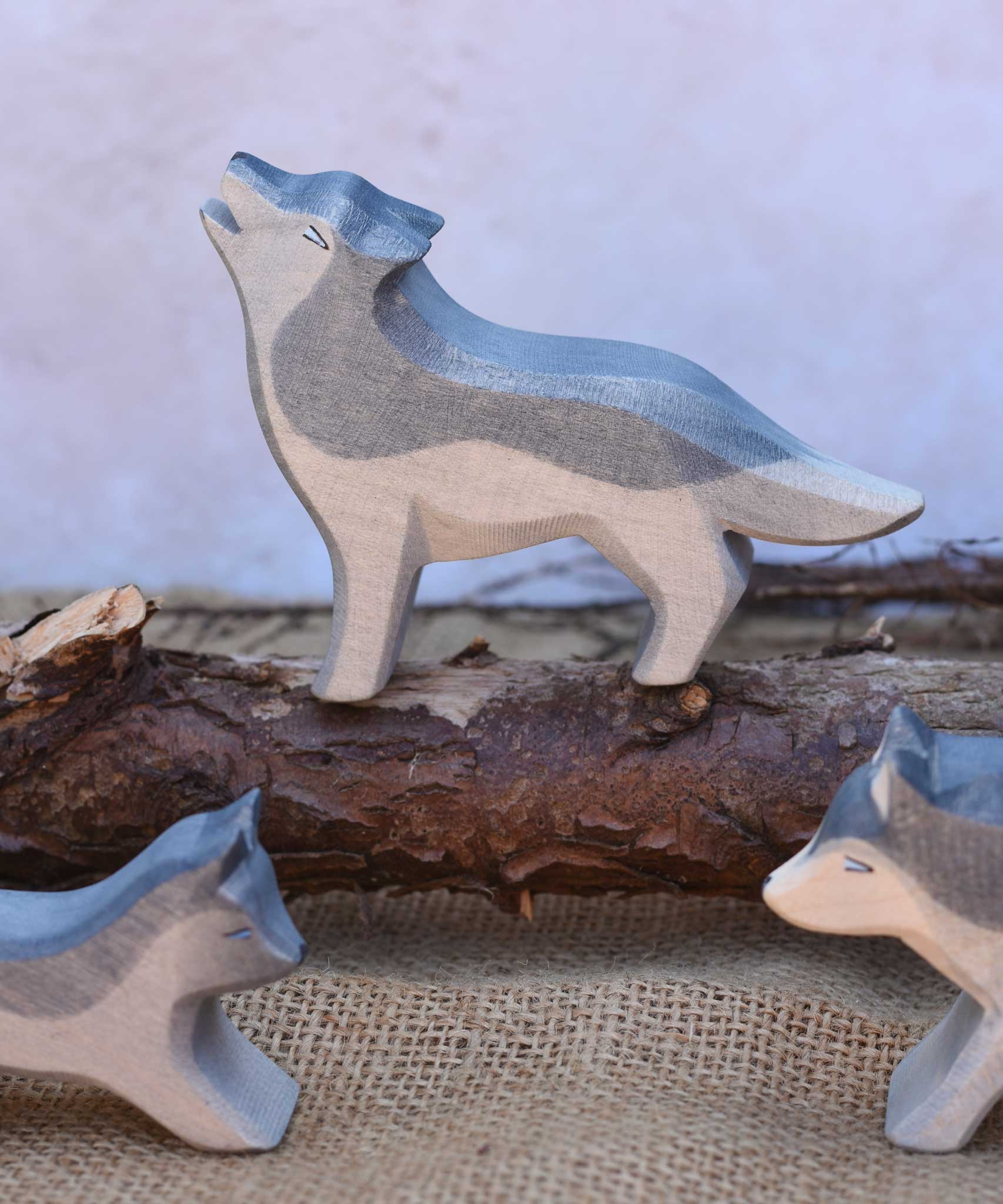 An Ostheimer Howling Wolf wooden toy figure shown in a small world scene set up with other Ostheimer wolves. The wolf is standing on a log branch. These figures are part of a vast range of expertly crafted wooden toy figures and scenery here at Babipur.