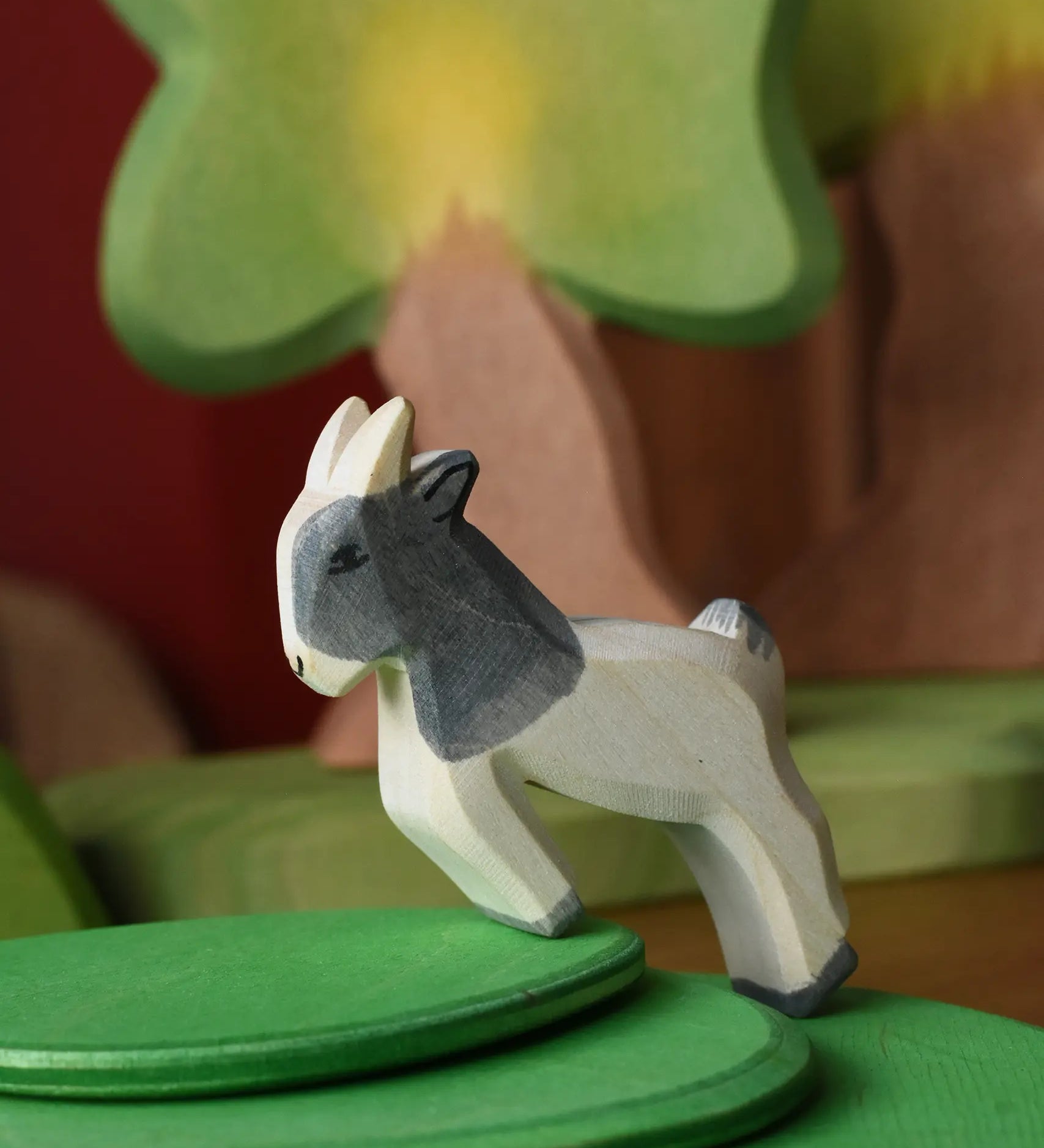Ostheimer white and grey jumping goat kid wooden toy figure on a stack of meadow plates with oak trees in the background. 