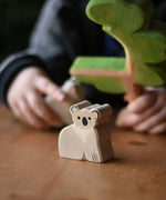 An Ostheimer koala placed on a wooden surface, a child can be seen in the bachground playing with a Ostheimer eucalyptus tree. These wooden figures are part of a wide range of expertly hand crafted wooden toys here at Babipur. 