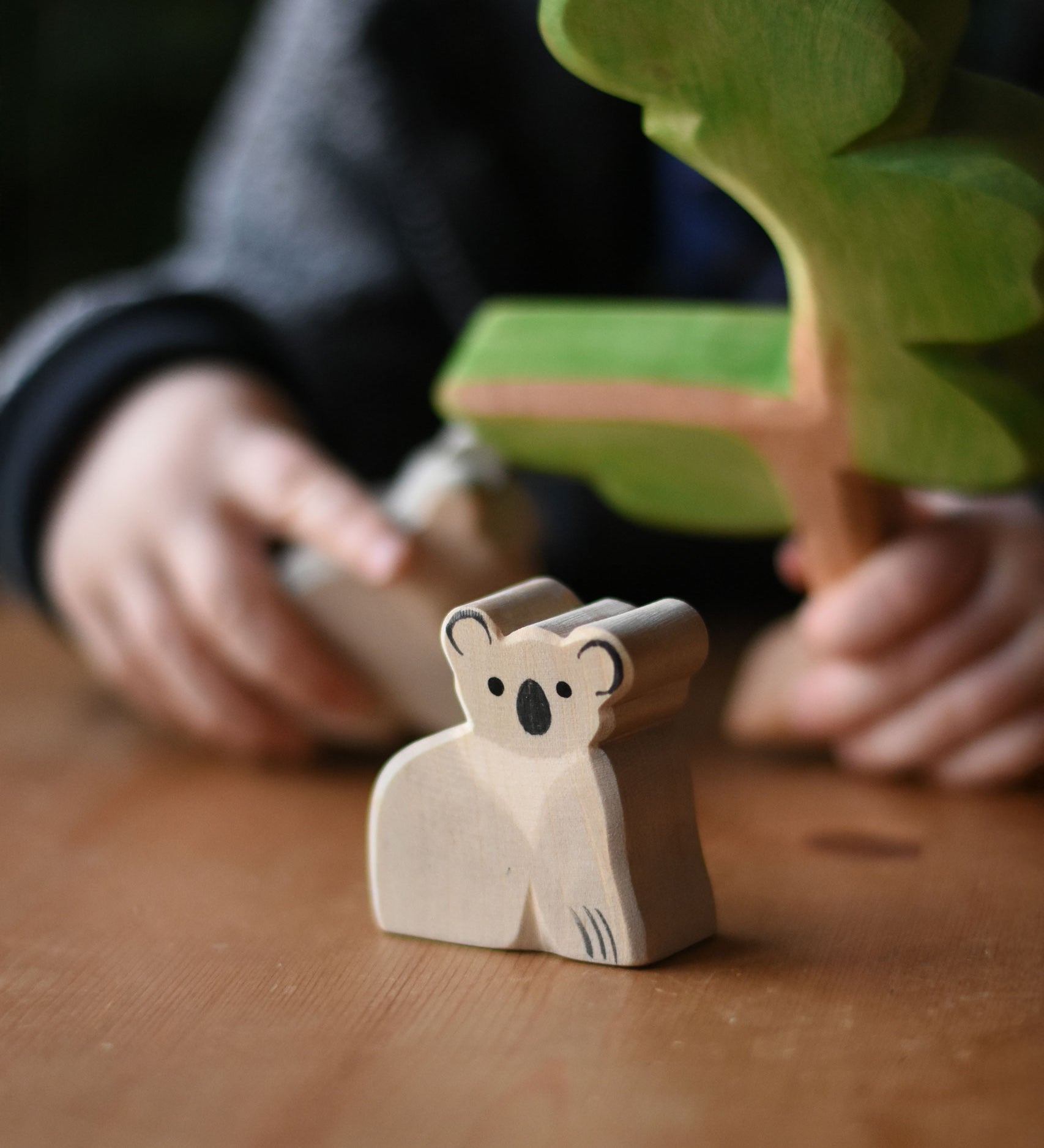 An Ostheimer koala placed on a wooden surface, a child can be seen in the bachground playing with a Ostheimer eucalyptus tree. These wooden figures are part of a wide range of expertly hand crafted wooden toys here at Babipur. 