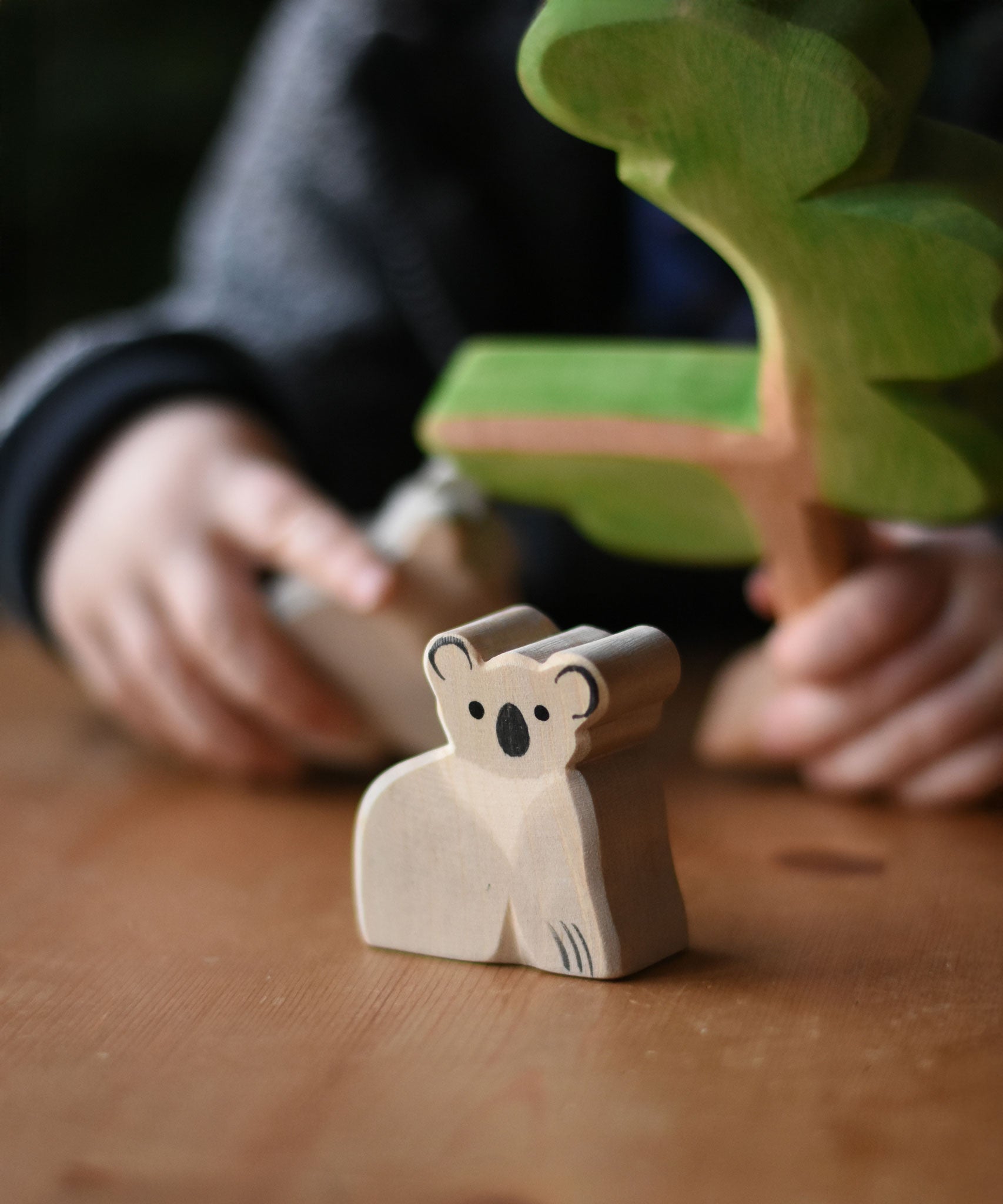 An Ostheimer koala placed on a wooden surface, a child can be seen in the bachground playing with a Ostheimer eucalyptus tree. These wooden figures are part of a wide range of expertly hand crafted wooden toys here at Babipur. 