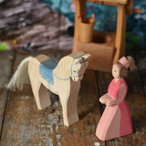 An Ostheimer hand made wooden Lady-In-Waiting toy figure and a horse with saddle placed on a wooden surface in a small world play scene. Other wooden toys including an Ostheimer water well can be seen in the background. These wooden figures are part of a wide range of expertly hand crafted wooden toys here at Babipur. 
