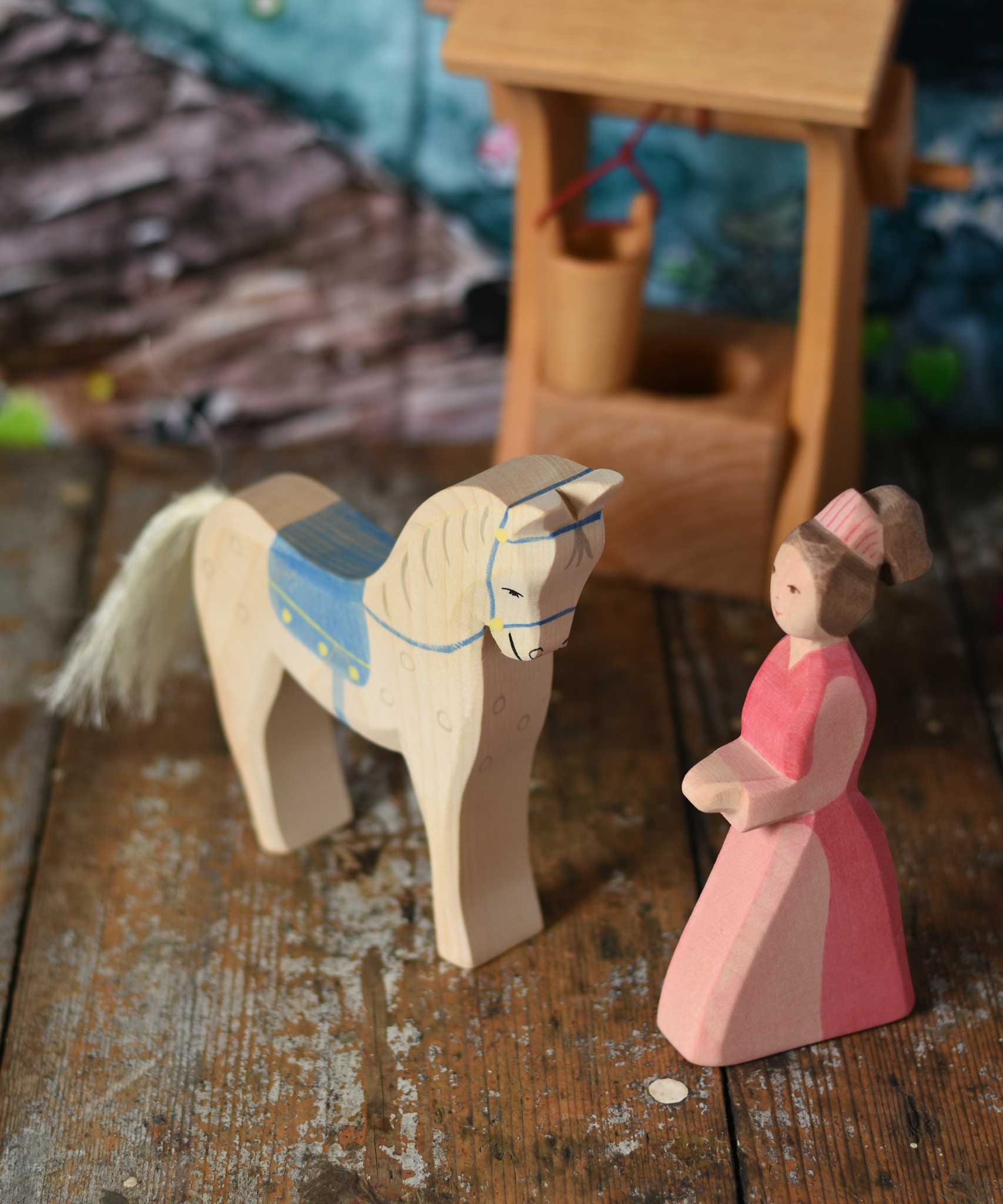 An Ostheimer hand made wooden Lady-In-Waiting toy figure and a horse with saddle placed on a wooden surface in a small world play scene. Other wooden toys including an Ostheimer water well can be seen in the background. These wooden figures are part of a wide range of expertly hand crafted wooden toys here at Babipur. 