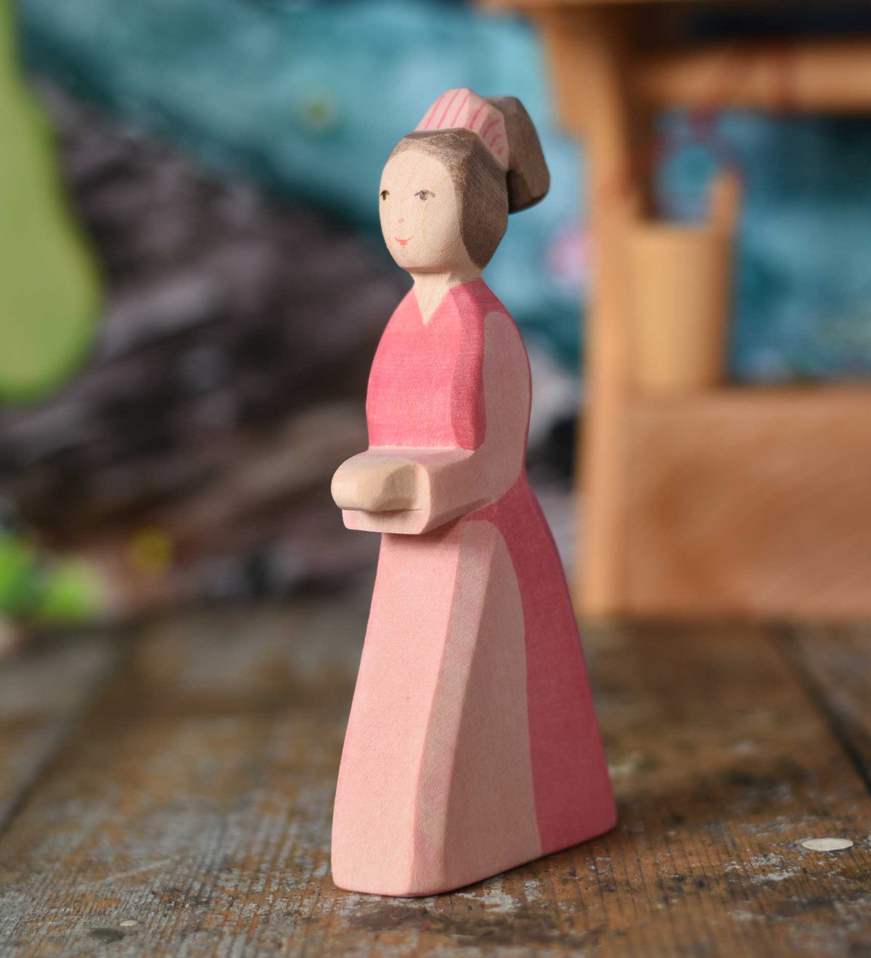 An Ostheimer hand made wooden Lady-In-Waiting toy figure placed on a wooden surface in a small world play scene. Other wooden toys including an Ostheimer water well can be seen in the background. These wooden figures are part of a wide range of expertly hand crafted wooden toys here at Babipur. 
