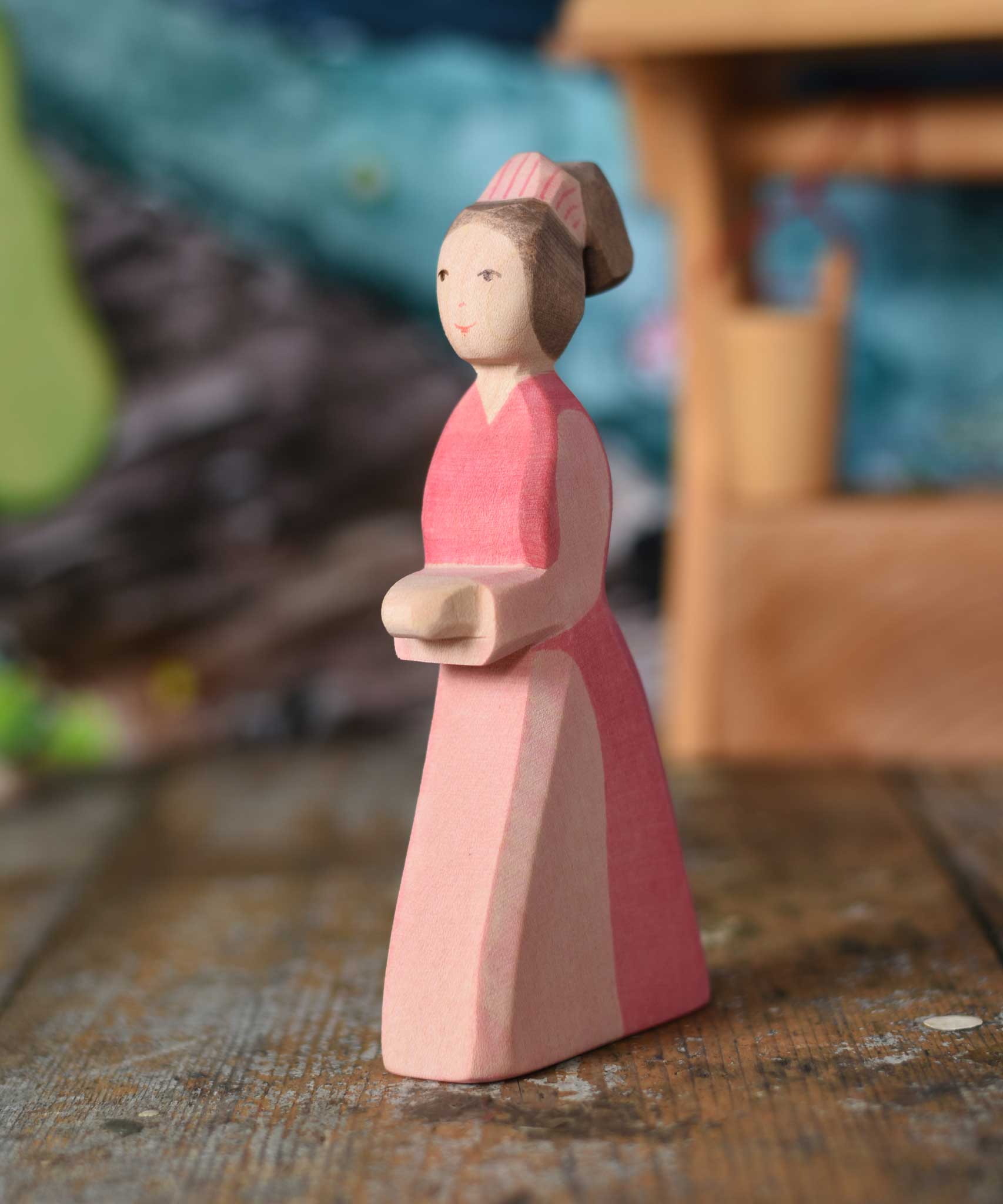 An Ostheimer hand made wooden Lady-In-Waiting toy figure placed on a wooden surface in a small world play scene. Other wooden toys including an Ostheimer water well can be seen in the background. These wooden figures are part of a wide range of expertly hand crafted wooden toys here at Babipur. 