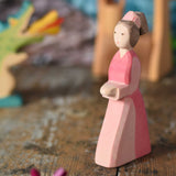 An Ostheimer hand made wooden Lady-In-Waiting toy figure placed on a wooden surface in a small world play scene. Other wooden toys including an Ostheimer water well can be seen in the background. These wooden figures are part of a wide range of expertly hand crafted wooden toys here at Babipur. 
