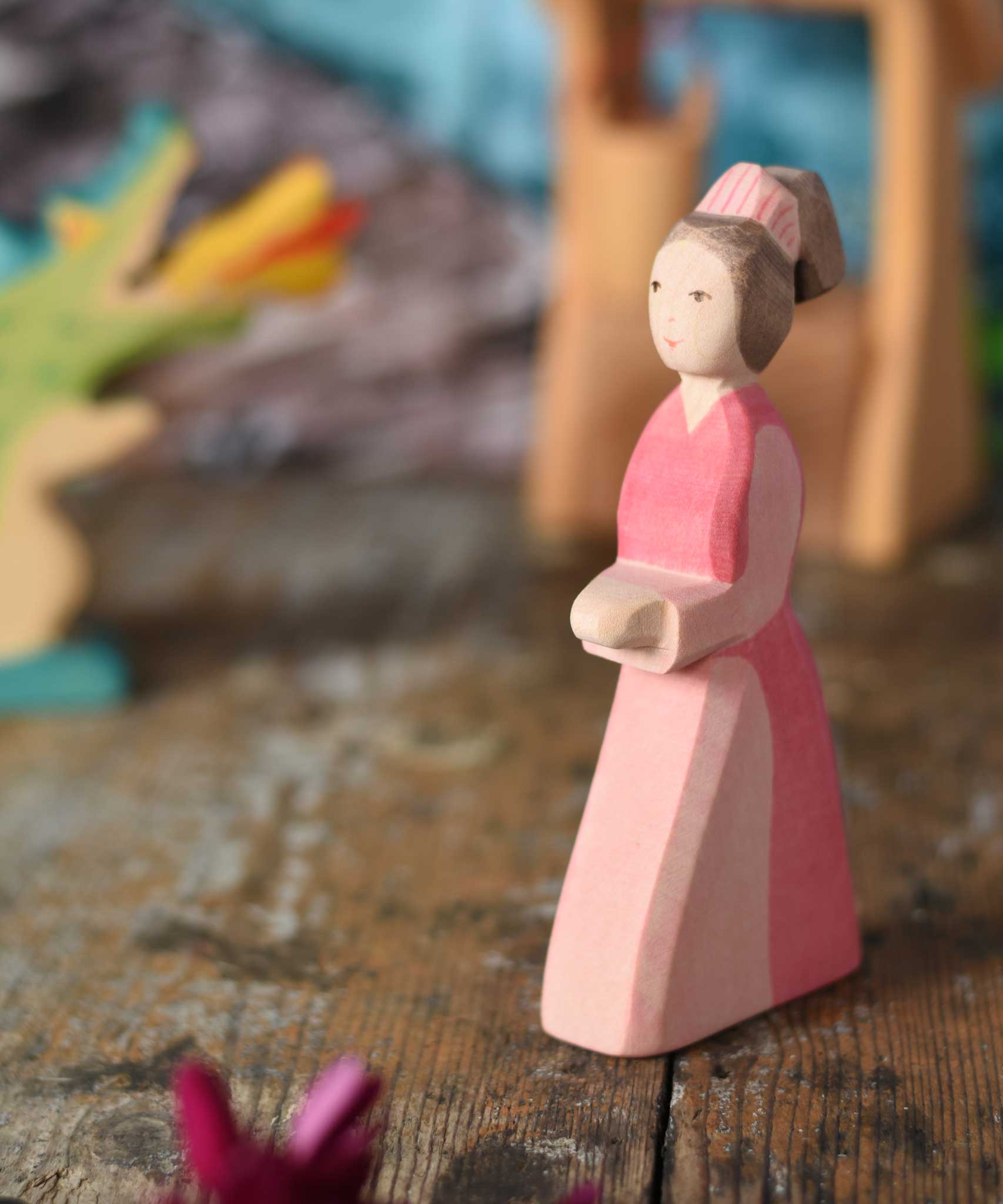 An Ostheimer hand made wooden Lady-In-Waiting toy figure placed on a wooden surface in a small world play scene. Other wooden toys including an Ostheimer water well can be seen in the background. These wooden figures are part of a wide range of expertly hand crafted wooden toys here at Babipur. 