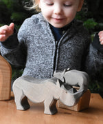 The large Ostheimer rhino wooden toy figure placed on a wooden surface. A child can be seen looking on excitedly in the background. These wooden figures are part of a wide range of expertly hand crafted wooden toys here at Babipur.