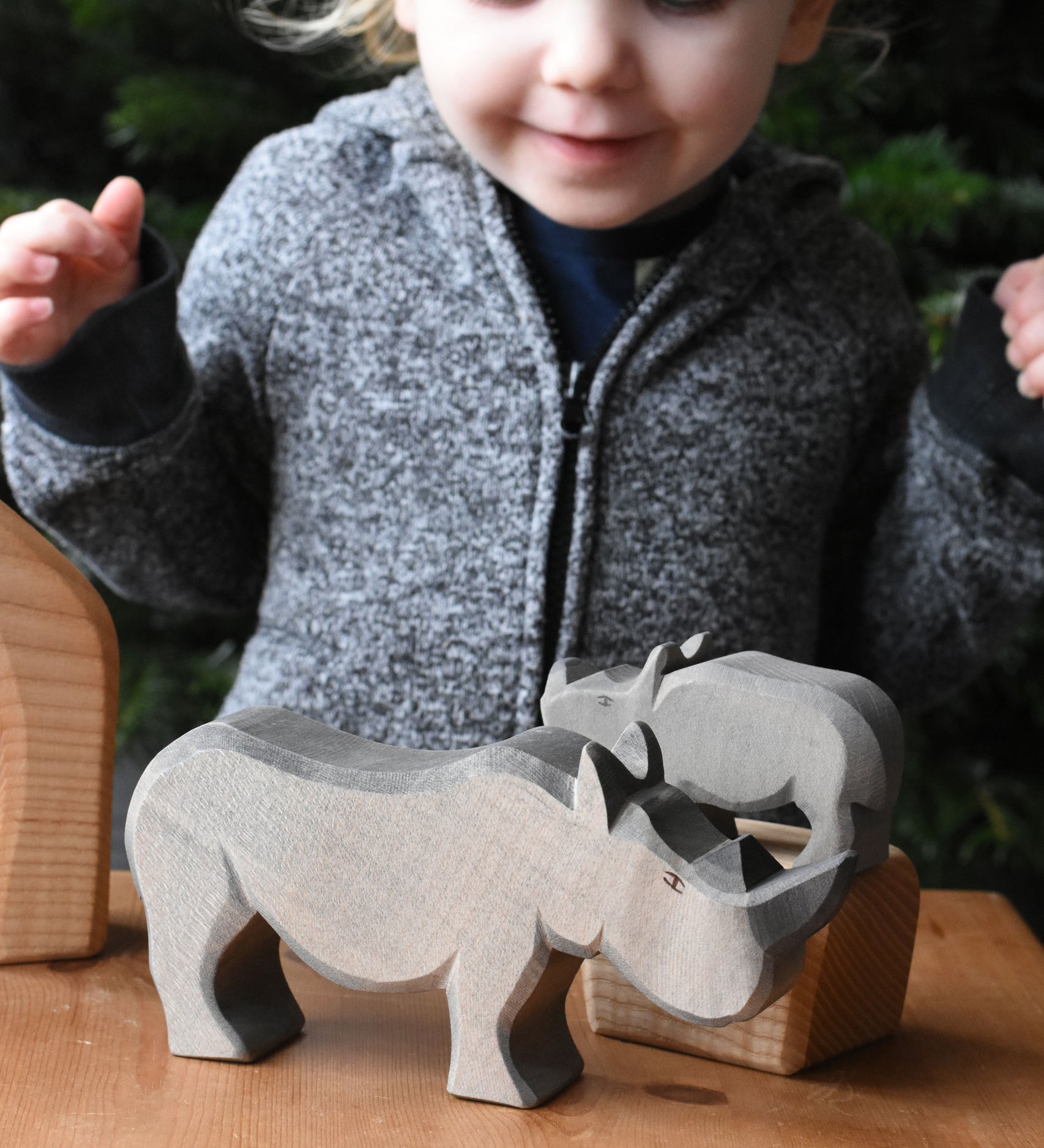 The large Ostheimer rhino wooden toy figure placed on a wooden surface. A child can be seen looking on excitedly in the background. These wooden figures are part of a wide range of expertly hand crafted wooden toys here at Babipur.