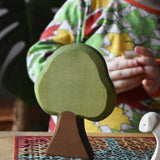 Ostheimer Linden wooden toy tree scenery piece for small world play placed on a rattan stool. Other Ostheimer toys and a child can be seen in the background. 