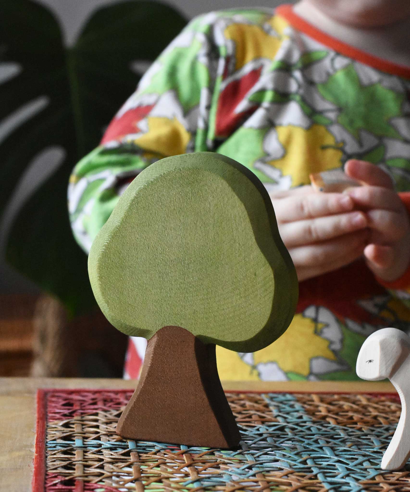 Ostheimer Linden wooden toy tree scenery piece for small world play placed on a rattan stool. Other Ostheimer toys and a child can be seen in the background. 
