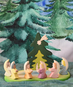 A host of little Ostheimer angels placed on a pond scene piece in a small world play set up. These little angel figures all have different coloured dresses, white, yellow, orange, purple, pink and blue with moon shaped wings. There is a Bumbu nativity tree puzzle behind them and a Wonderie Christmas tree WonderCloth hung up in the back. These wooden figures are part of a wide range of expertly hand crafted wooden toys here at Babipur. 