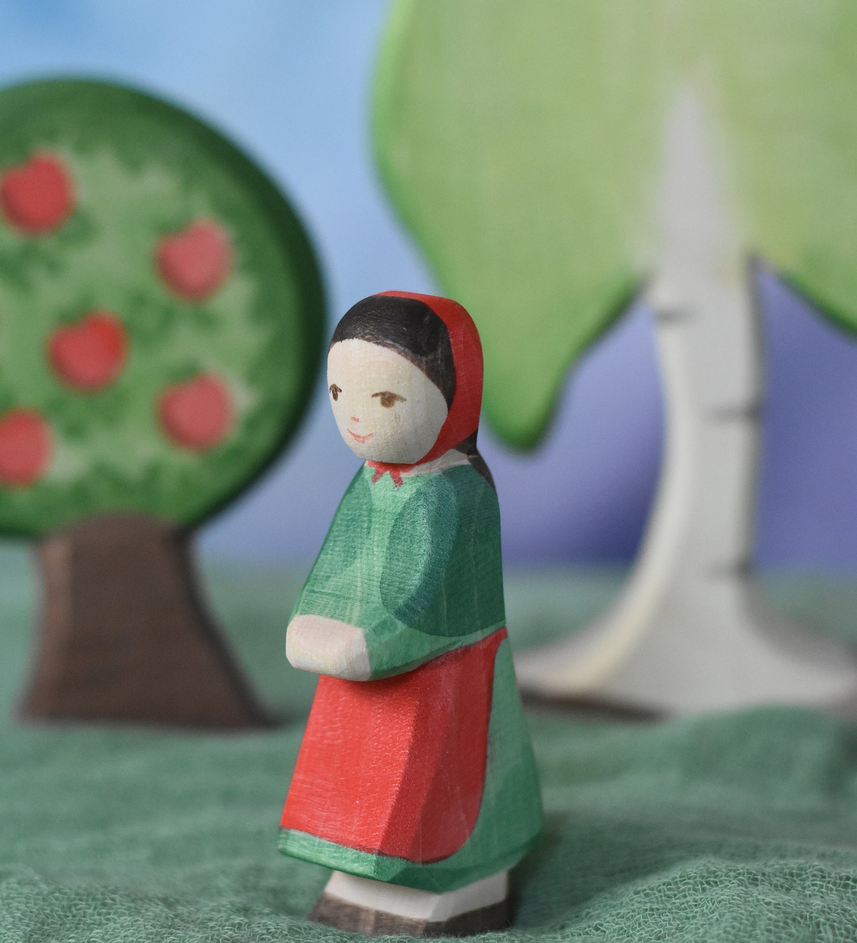 An Ostheimer wooden little red riding hood toy figure placed in the middle of a small world play scene. There is an apple tree and large birch tree in the background with a green coloured decorative scarf underneath them. These wooden figures are part of a wide range of expertly hand crafted wooden toys here at Babipur. 