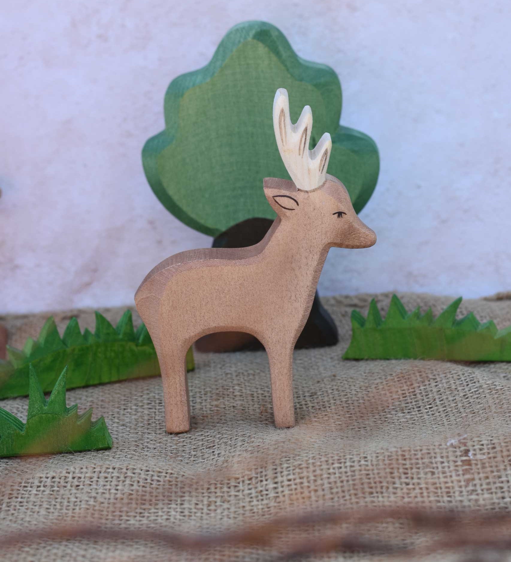 An Ostheimer male deer toy figure placed in the middle of a small world play scene. Ostheimer grass an oak tree figure can be seen in the background. Check out our full range of expertly crafted wooden toy figures and scenery here at Babipur.