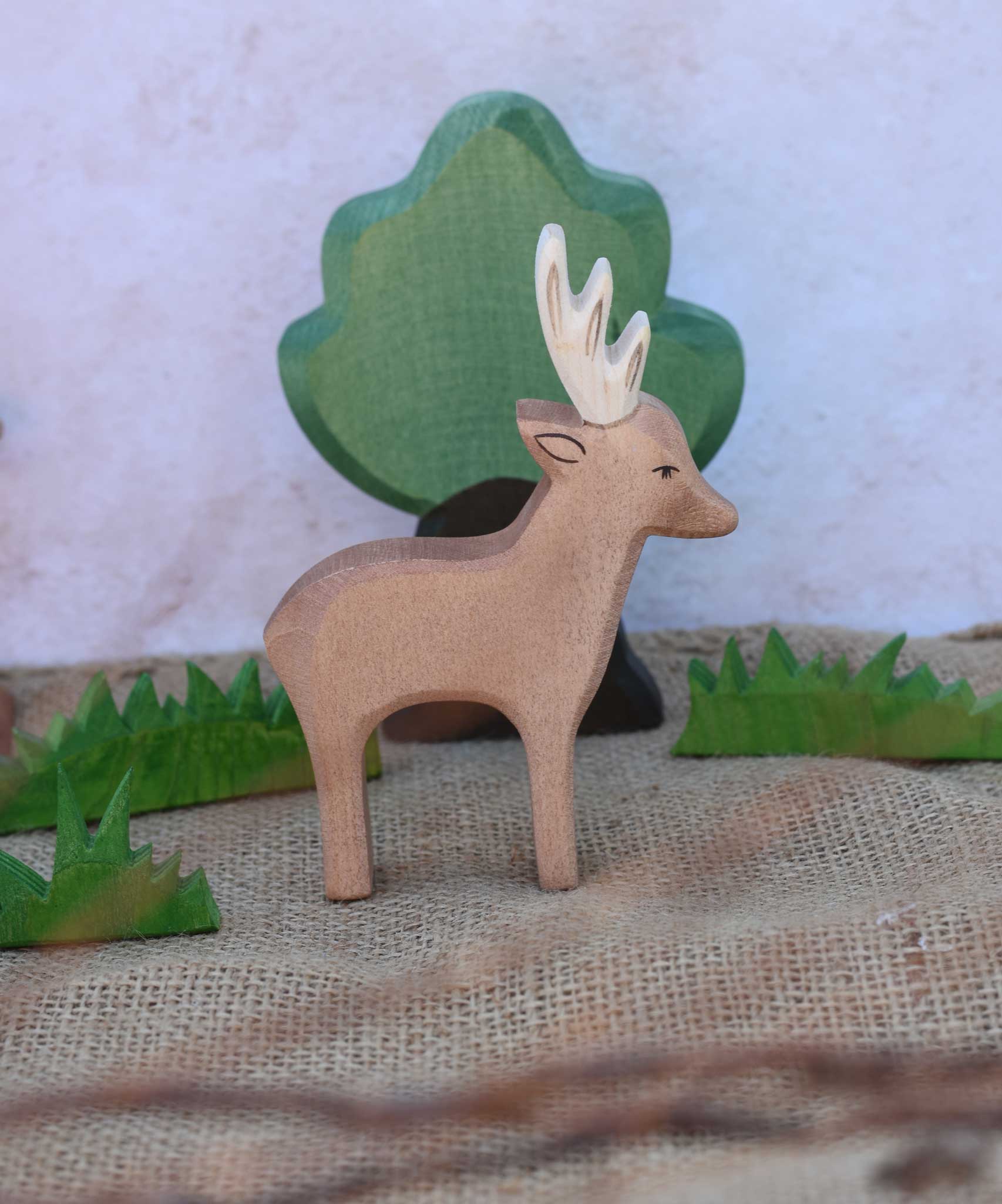 An Ostheimer male deer toy figure placed in the middle of a small world play scene. Ostheimer grass an oak tree figure can be seen in the background. Check out our full range of expertly crafted wooden toy figures and scenery here at Babipur.
