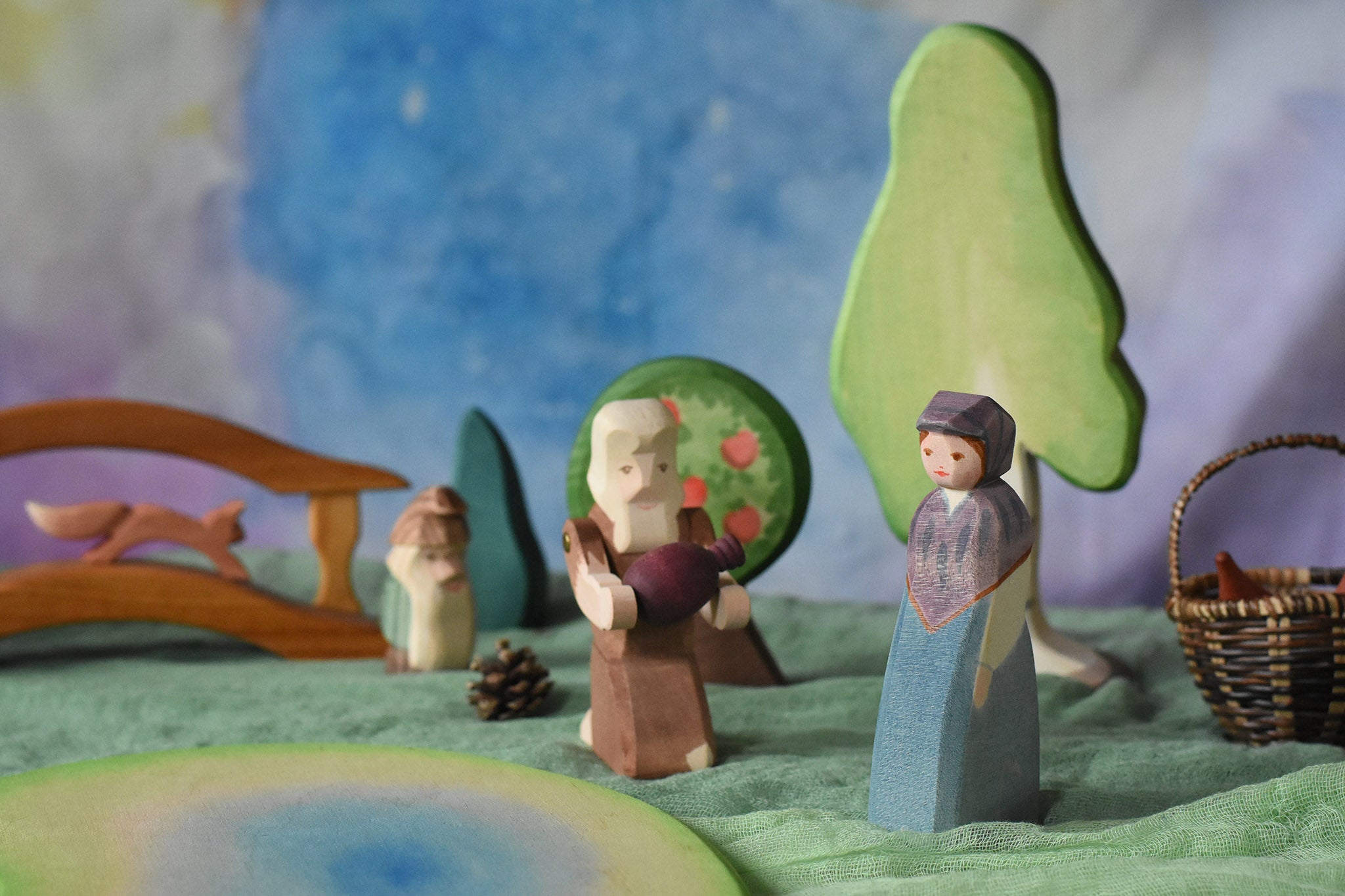An Ostheimer market woman wooden toy figure placed in a small world play scene. The Ostheimer Beggar figure can be seen in the background along with scenery pieces such as trees and a wooden bridge. This wooden figure is part of a large range of expertly hand crafted, Waldorf inspired, wooden toys here at Babipur. 
