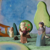 An Ostheimer market woman wooden toy figure placed in a small world play scene. The Ostheimer Beggar figure can be seen in the background along with scenery pieces such as trees and a wooden bridge. This wooden figure is part of a large range of expertly hand crafted, Waldorf inspired, wooden toys here at Babipur. 