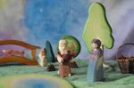 An Ostheimer market woman wooden toy figure placed in a small world play scene. The Ostheimer Beggar figure can be seen in the background along with scenery pieces such as trees and a wooden bridge. This wooden figure is part of a large range of expertly hand crafted, Waldorf inspired, wooden toys here at Babipur. 