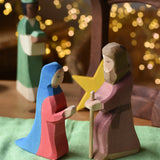 Ostheimer Mary and Joseph wooden toy figures in a festive small world play scene with fairy lights in the background.