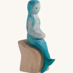 A wooden Ostheimer mermaid with blue hair sitting on a rock and on a cream background.