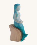 A wooden Ostheimer mermaid with blue hair sitting on a rock and on a cream background.