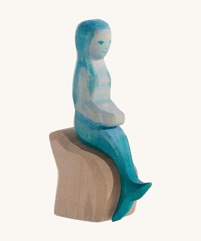 A wooden Ostheimer mermaid with blue hair sitting on a rock and on a cream background.