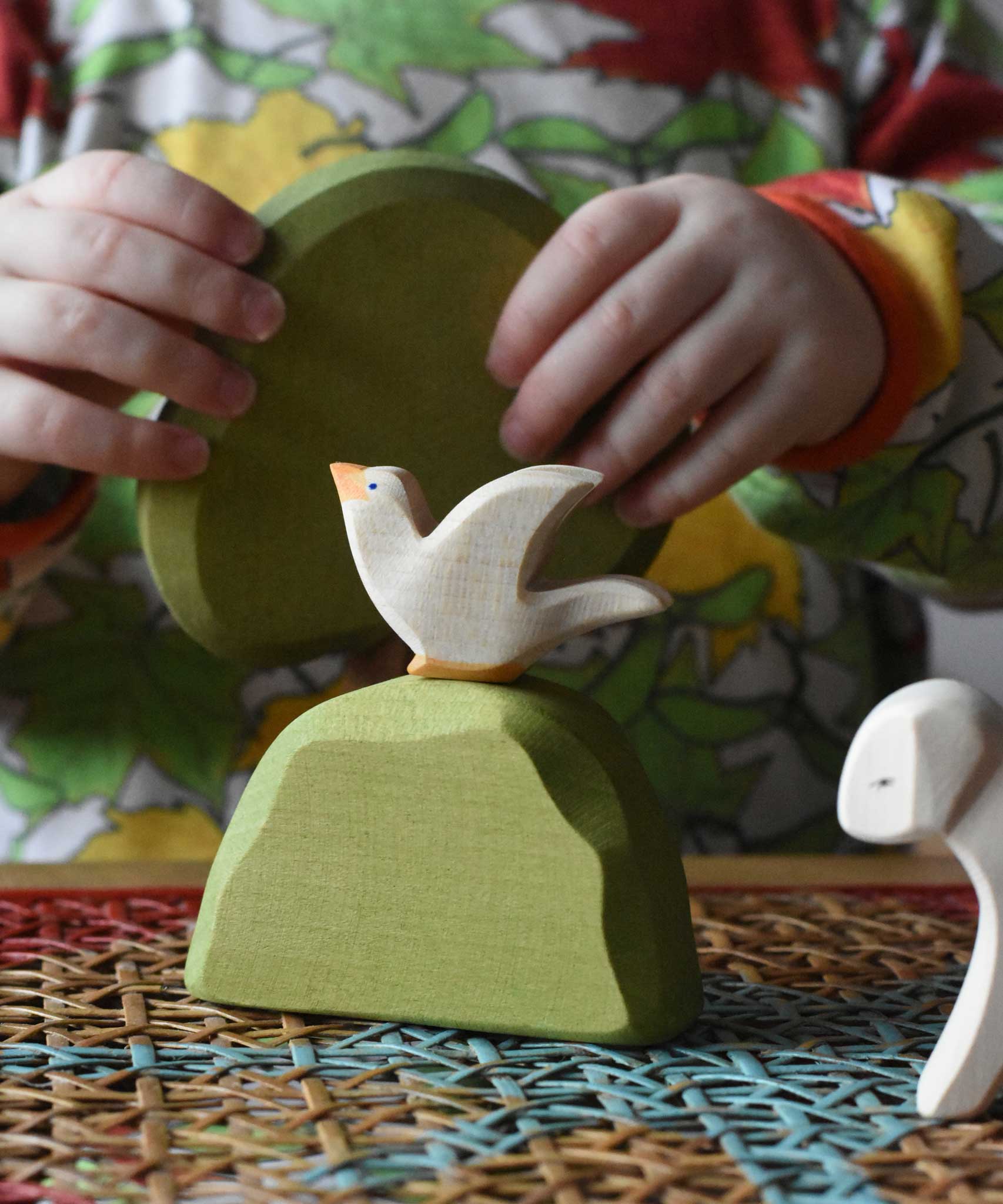 Ostheimer white dove bird figure placed on a middle green bush on a rattan top stool placed in a simple small world play scene. 