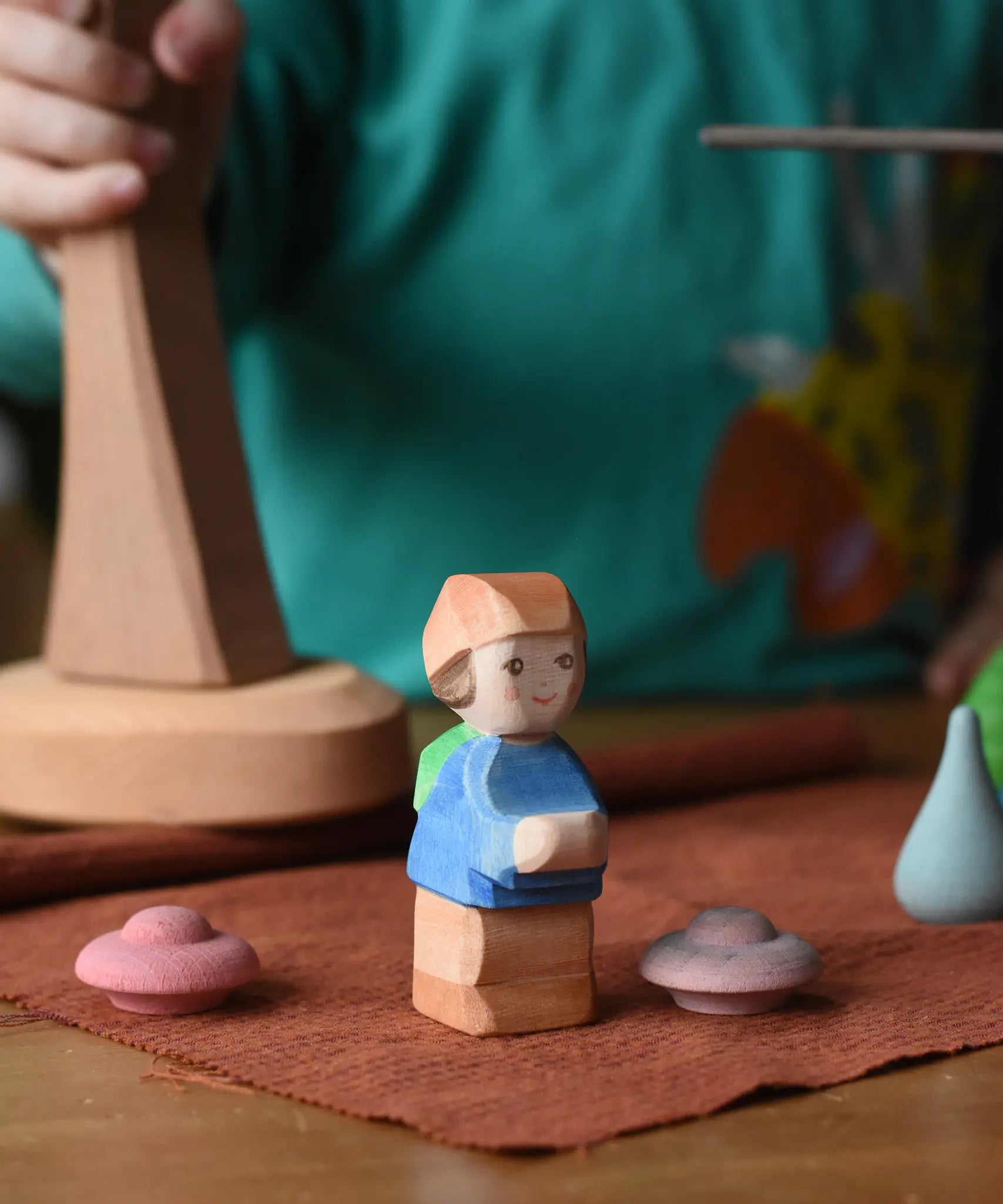 Ostheimer Mo handmade little boy wooden toy figure placed on a brown cloth with pink Grapat mandala pieces around it.