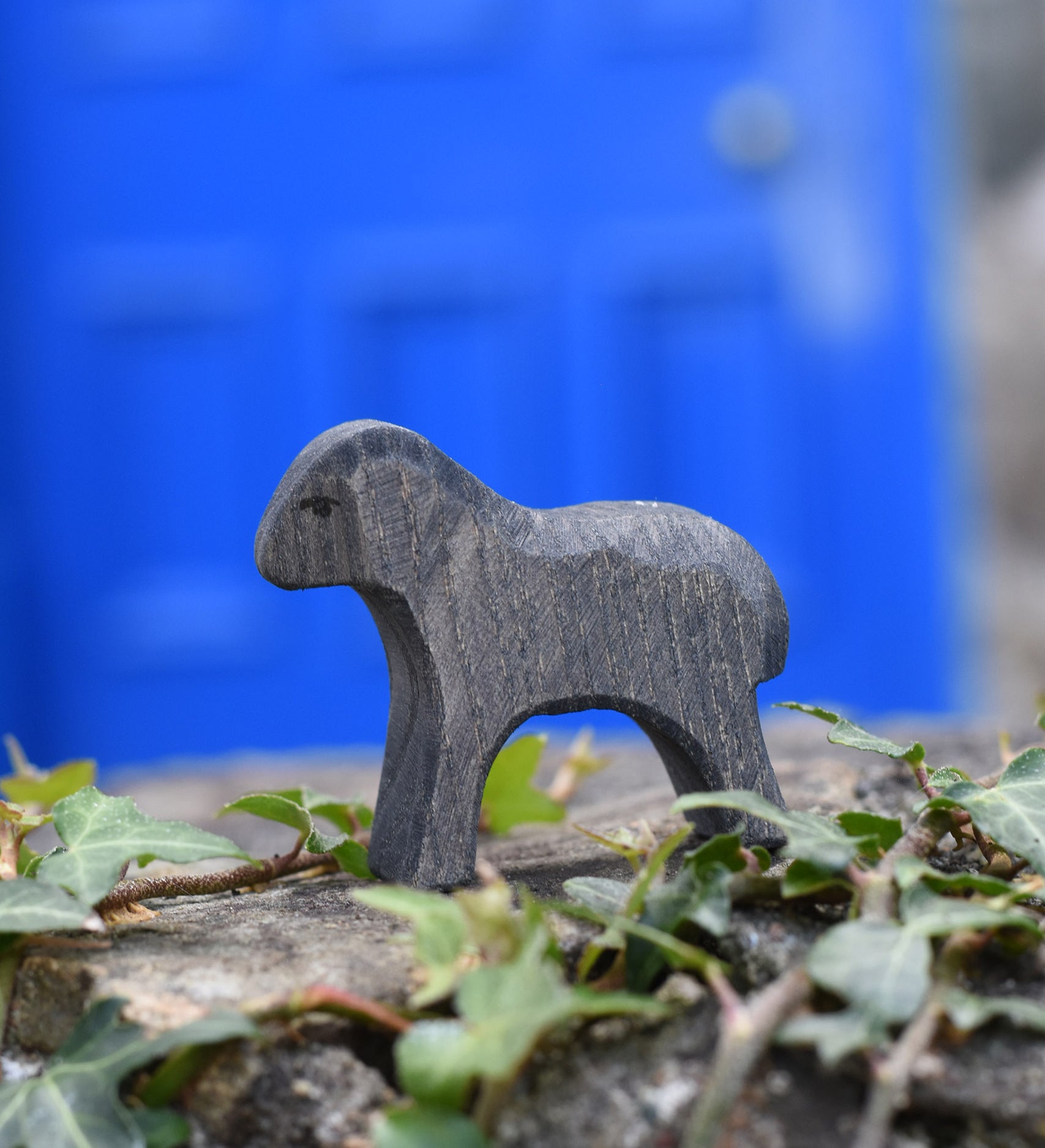 An Ostheimer wooden Moorland lamb figure placed on a stone wall outdoors. These wooden figures are part of a large range of expertly hand crafted, Waldorf inspired, wooden toys here at Babipur.