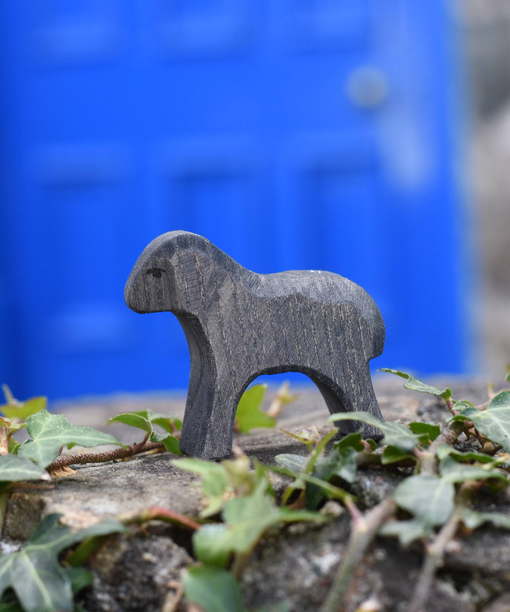 An Ostheimer wooden Moorland lamb figure placed on a stone wall outdoors. These wooden figures are part of a large range of expertly hand crafted, Waldorf inspired, wooden toys here at Babipur.