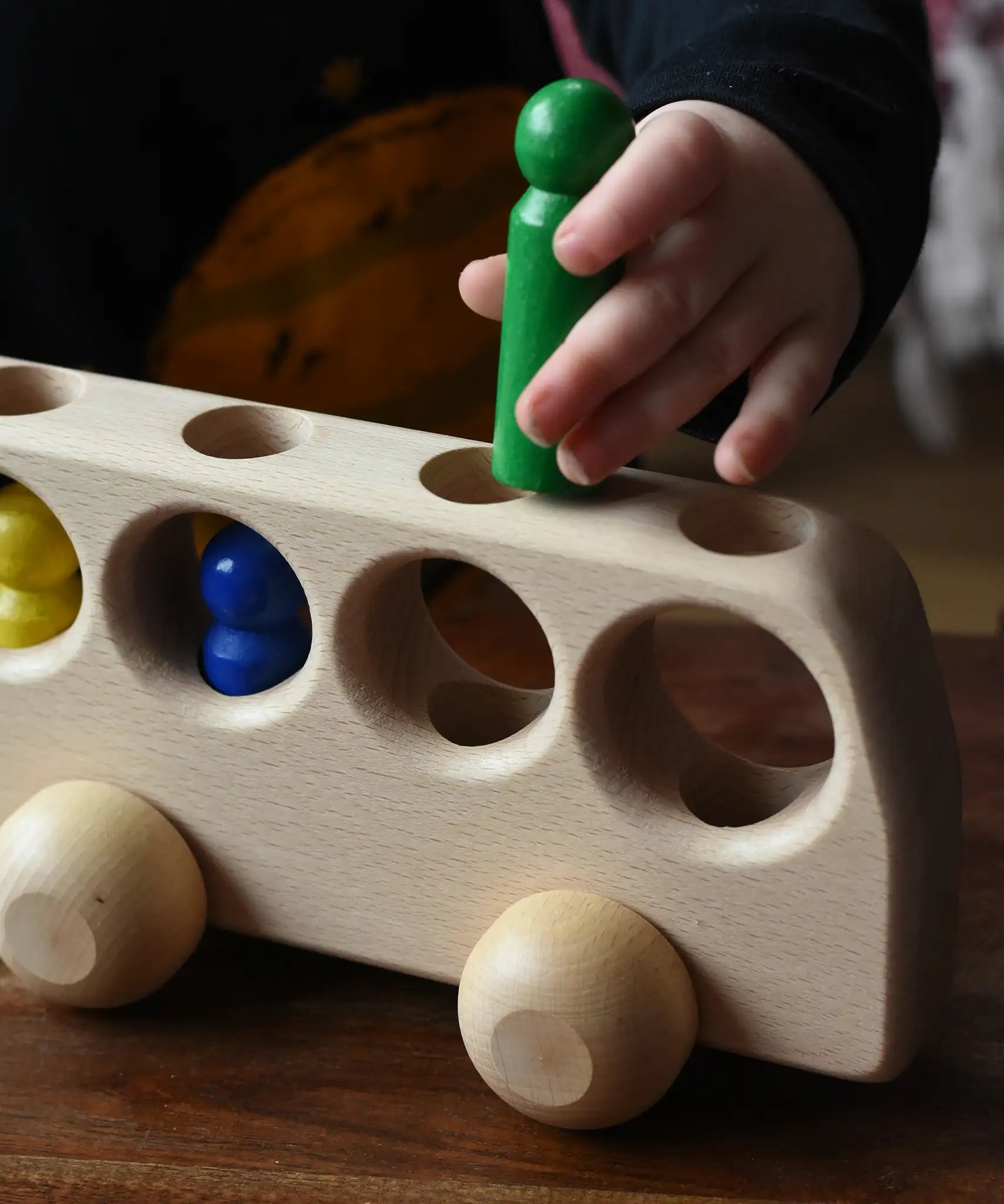Close up of a child playing with the Ostheimer Natural Bus With 4 People. The child is placing the green coloured wooden figures into one of the circular shaped slots on the top of the bus. 