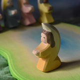 An orange coloured little angel figure placed on a wooden pond scene piece. Other little angels can be seen in the background.