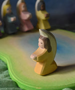 An orange coloured little angel figure placed on a wooden pond scene piece. Other little angels can be seen in the background.
