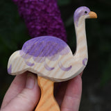 An Ostheimer ostrich shown in an adult's hand for scale. Purple buddleia flowers can be seen in the background. 