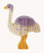 A side view of Ostheimer Ostrich wooden toy figure. The photo shows the natural wood grain detail on the handcrafted figure. The ostrich has a white base, light yellow orange legs and beak and purple coloured details.