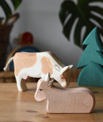 Ostheimer ox wooden toy figure placed on a wooden surface with a small spruce tree and standing ox in the background. 