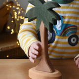 Ostheimer palm tree wooden toy figure with support base placed on a wooden surface. Child's hand reaching for the tree. 