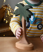 Ostheimer palm tree wooden toy figure with support base placed on a wooden surface. Child's hand reaching for the tree. 