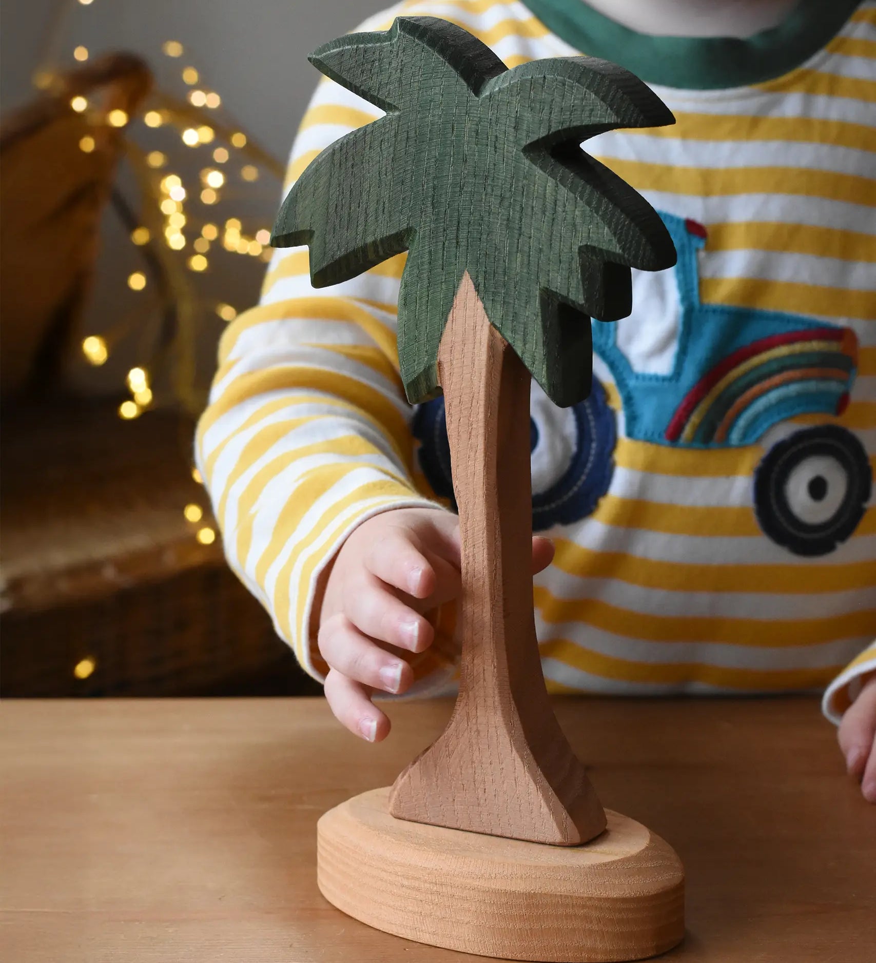 Ostheimer palm tree wooden toy figure with support base placed on a wooden surface. Child's hand reaching for the tree. 