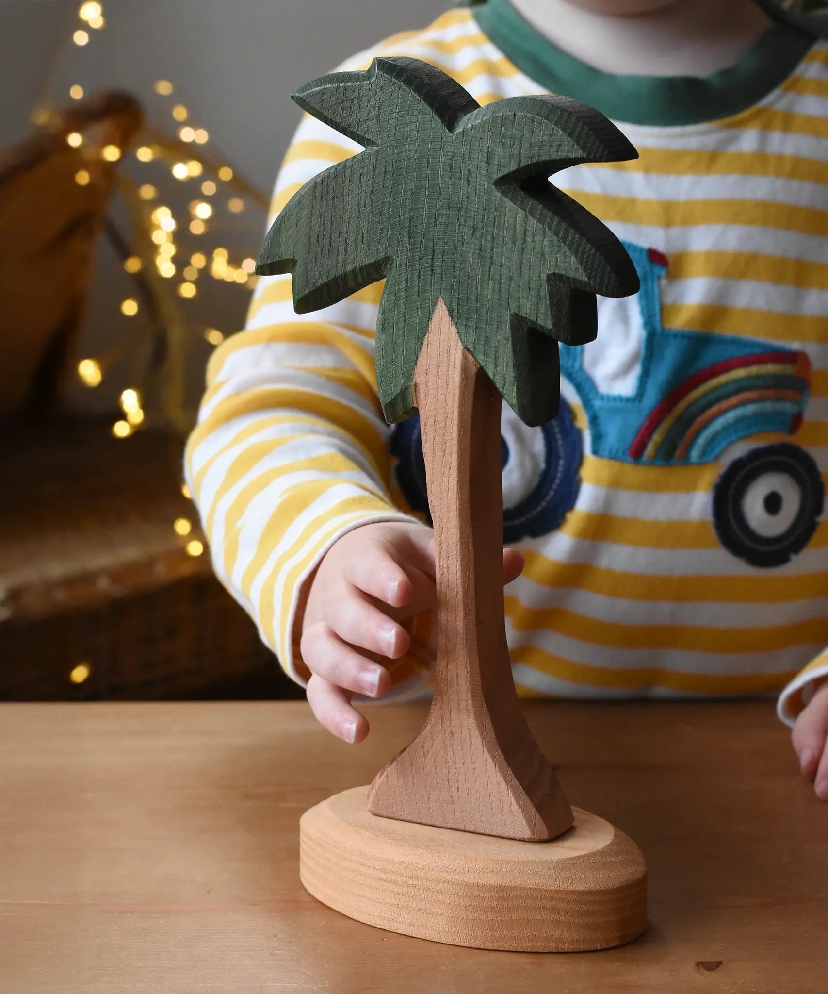 Ostheimer palm tree wooden toy figure with support base placed on a wooden surface. Child's hand reaching for the tree. 