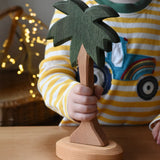Ostheimer palm tree wooden toy figure with support base placed on a wooden surface. Child holding the trunk.