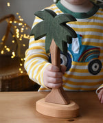 Ostheimer palm tree wooden toy figure with support base placed on a wooden surface. Child holding the trunk.