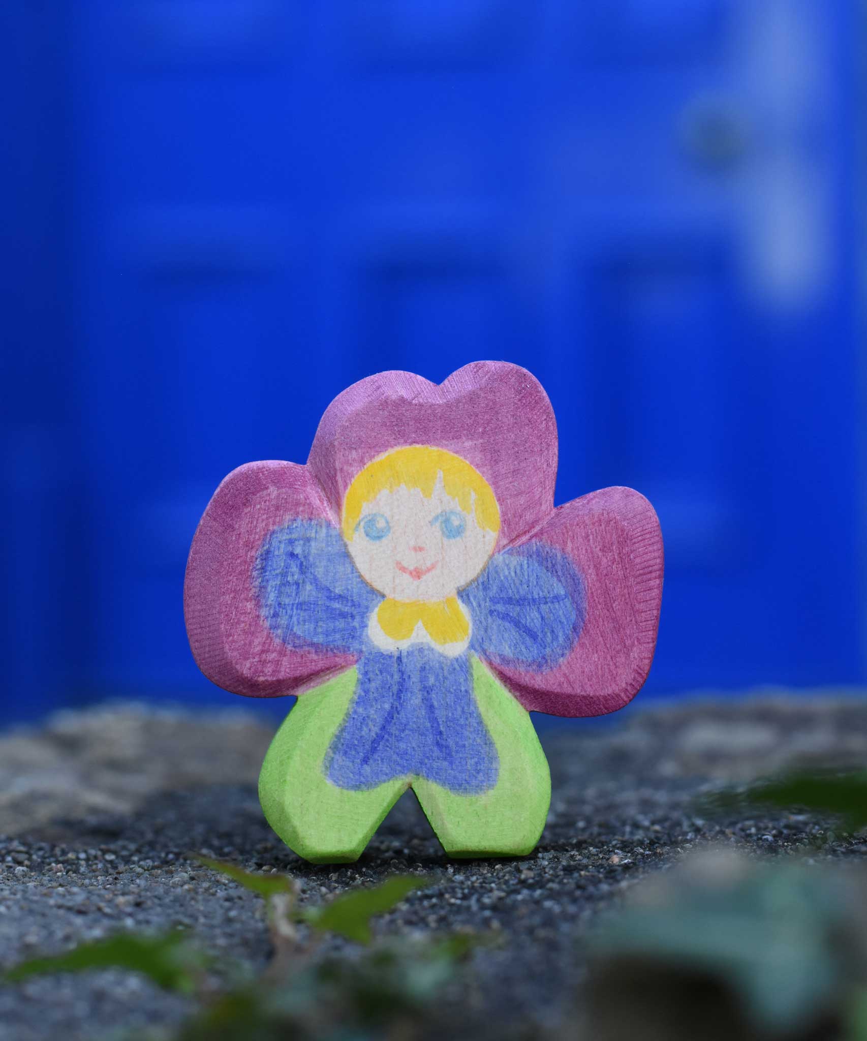 An Ostheimer hand made and hand painted wooden pansy Flower Child figure toy placed on a stone wall outdoors. These flower child wooden figures are part of a wide range of expertly hand crafted wooden toys here at Babipur. 