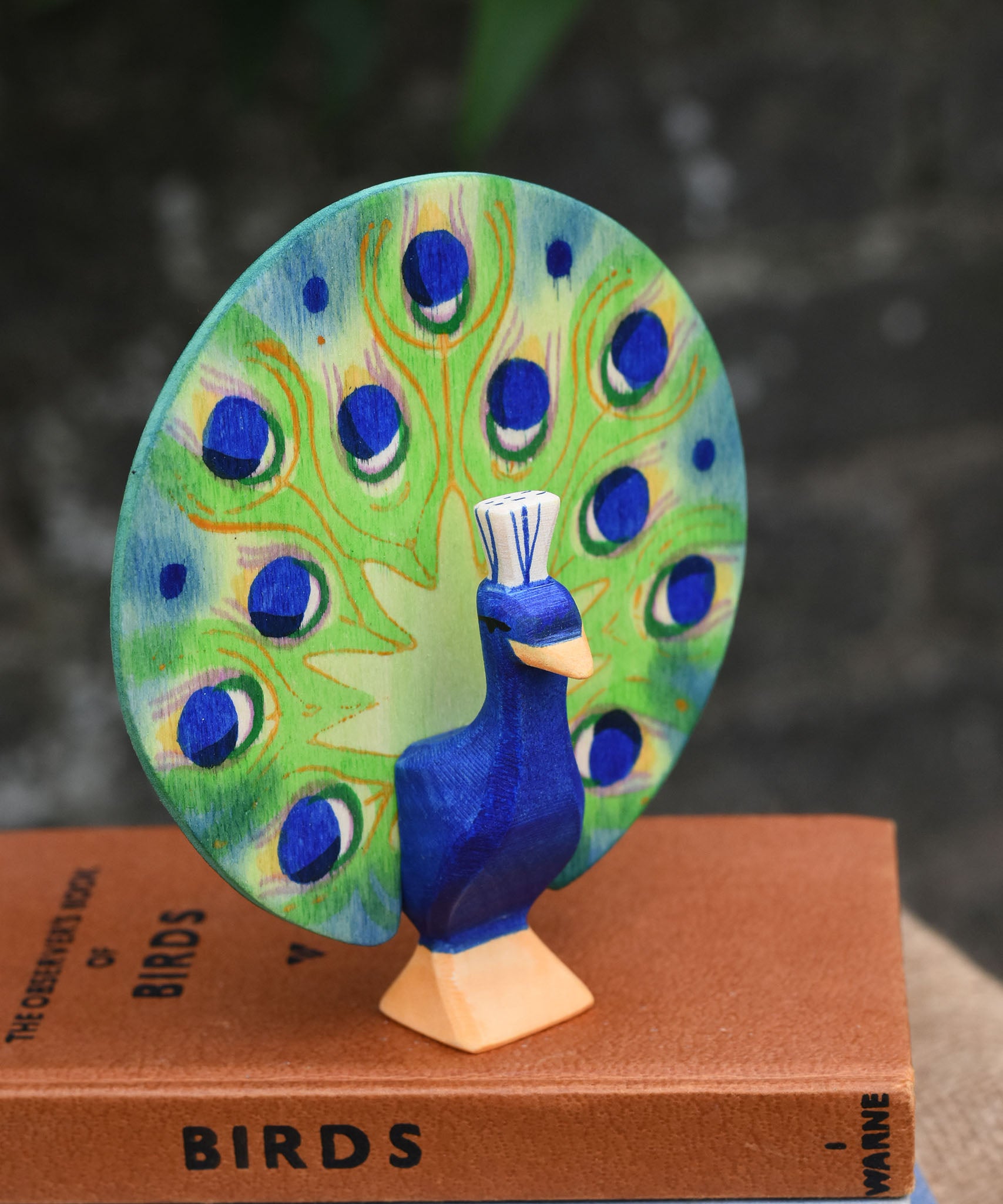 A Ostheimer peacock wooden toy figure with open feathers placed on a pile of books.