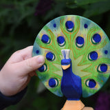 A close up of a child's hand holding the Ostheimer peacock wooden toy figure with open feathers.
