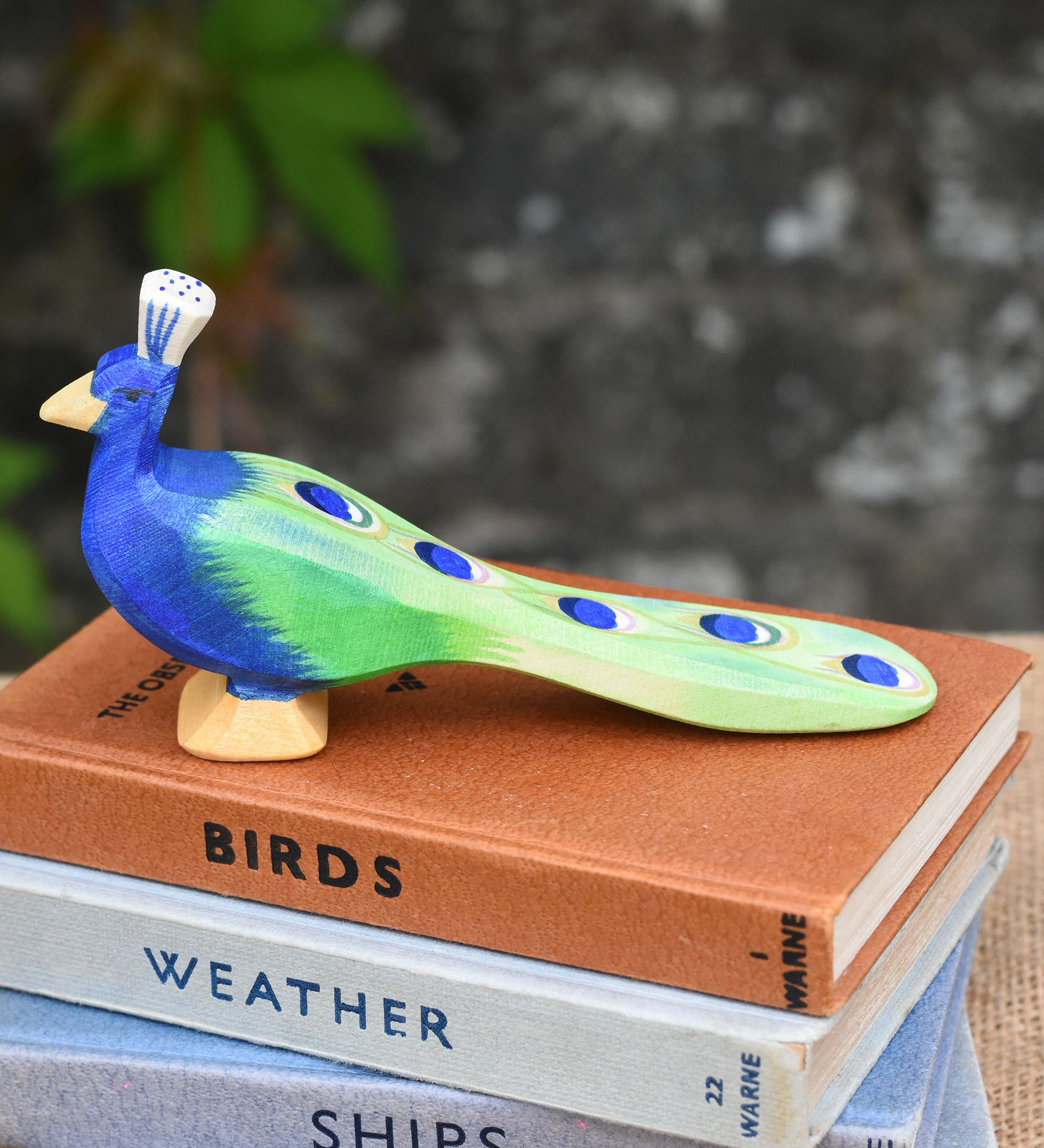 An Ostheimer peacock wooden toy figure with it's feathersdown  placed on a pile of books.