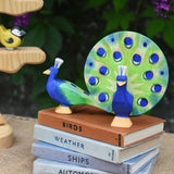 A Ostheimer peacock wooden toy figure with open feathers and another with feathers down placed on a pile of books with the Ostheimer bird tree in the background.