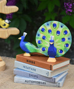 A Ostheimer peacock wooden toy figure with open feathers and another with feathers down placed on a pile of books with the Ostheimer bird tree in the background.