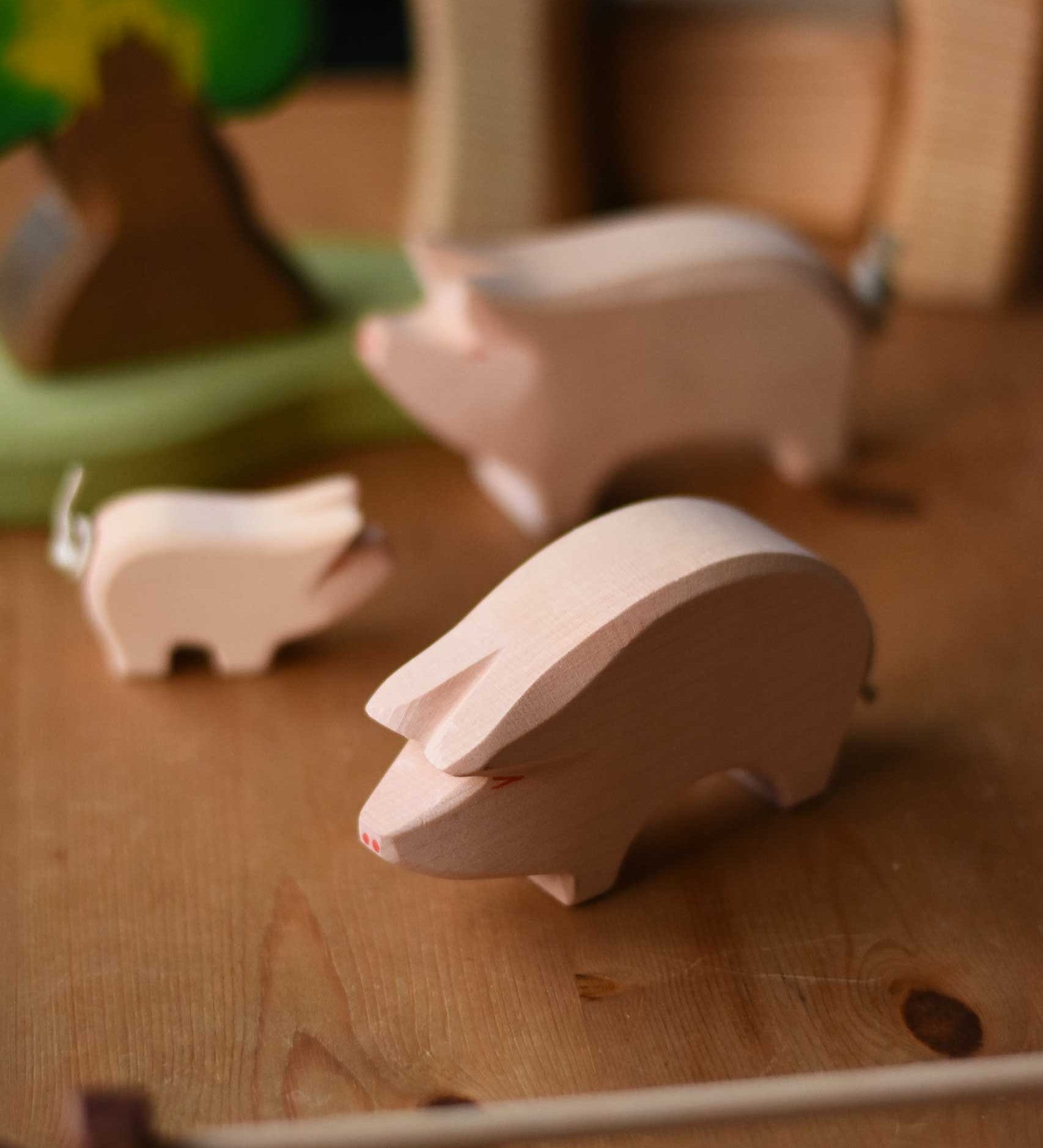 Ostheimer Pig with it's Head low placed on a wooden surface in a small world play with other pigs in the background. This pink pig has a little curled rope tail.  These wooden figures are part of a wide range of expertly hand crafted wooden toys here at Babipur. 