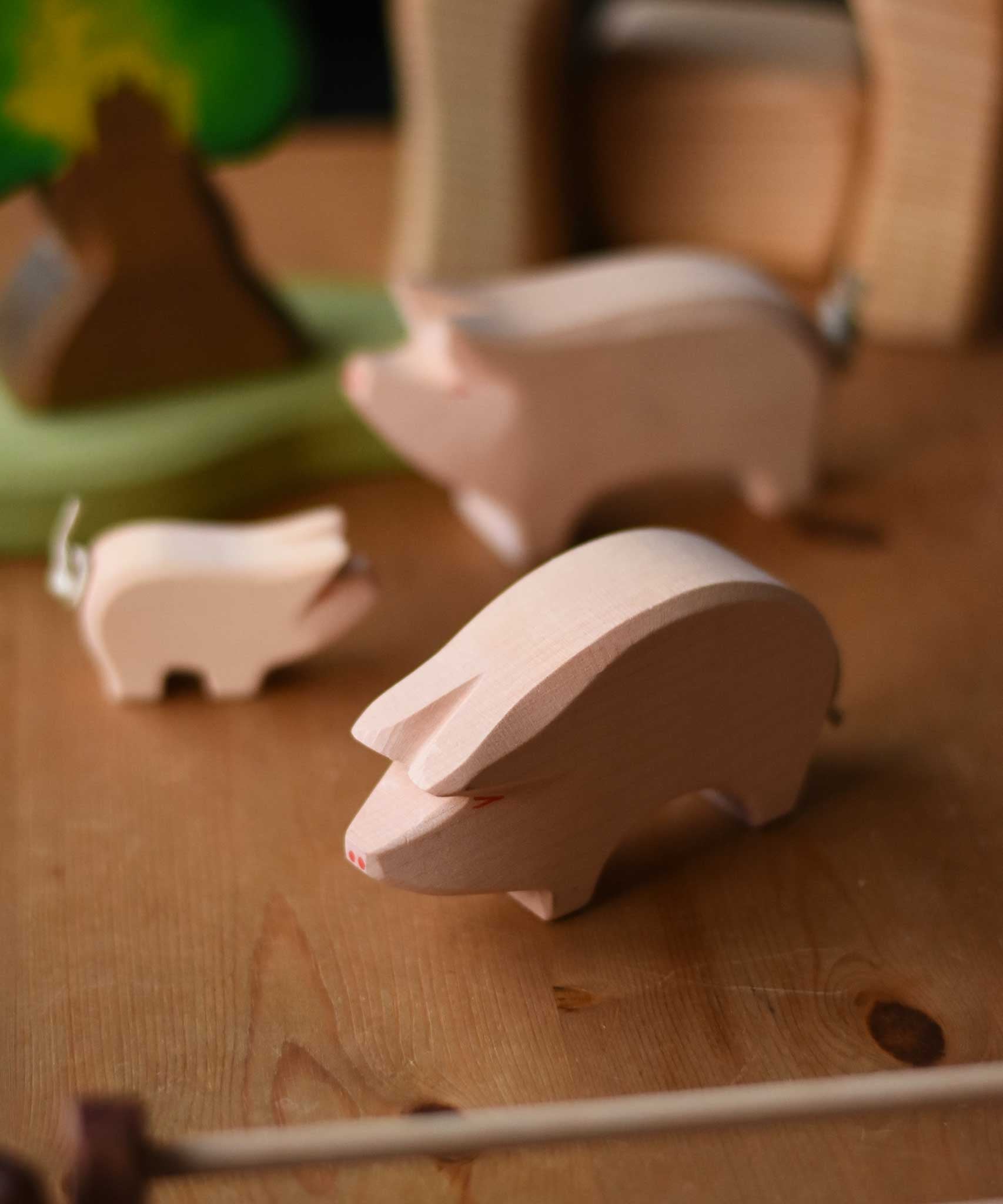 Ostheimer Pig with it's Head low placed on a wooden surface in a small world play with other pigs in the background. This pink pig has a little curled rope tail.  These wooden figures are part of a wide range of expertly hand crafted wooden toys here at Babipur. 